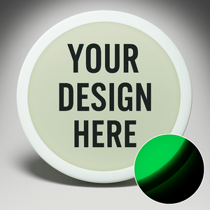 A custom glow-in-the-dark disc golf disc with the text 'YOUR DESIGN HERE' displayed on it. The disc appears white with a green glow effect on the text and a small inset image showing a close-up of the glow texture.